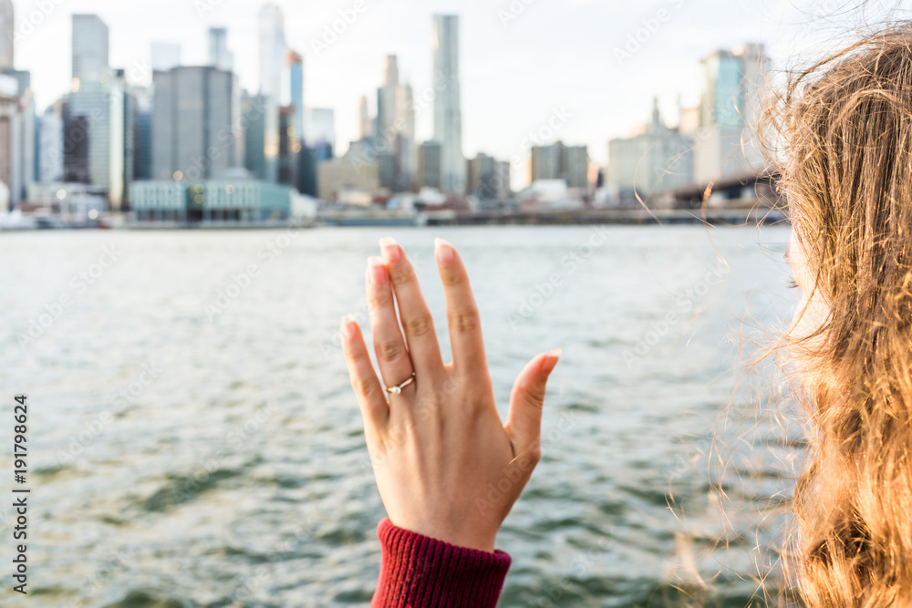 Young woman's hand with diamond engagement ring solitaire, gold wedding band outside outdoors in NYC New York City Brooklyn Bridge Park by east river, cityscape, skyline waving goodbye