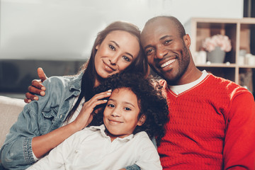 Portrait of happy mother, father and daughter sitting together at home, looking at camera and smiling