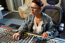 Thoughtful Modern Female Audio Engineer Concentrated On Mixing Sounds While Moving Faders And Twisting SSL Channels On Digital Audio Channel Mixer In Studio