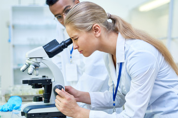 Wall Mural - Side view portrait of young female scientist looking in microscope while working on medical research in science laboratory