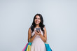 © MARIIA - Online shopping. Beautiful young woman holding shopping bags and using her smartphone with smile while standing against white background