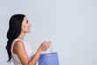 © MARIIA - Maybe this can solve my problems? Side view of beautiful young woman looking away and holding shopping bag with smile while standing against white background