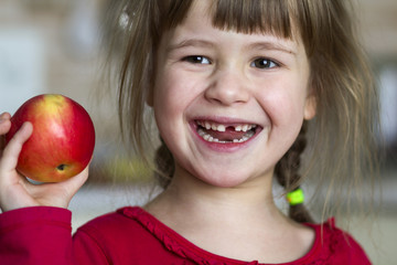 Poster - A cute little curly toothless girl smiles and holds a red apple. Portrait of a happy baby eating a red apple. The child loses milk teeth. Healthy food nutrition.