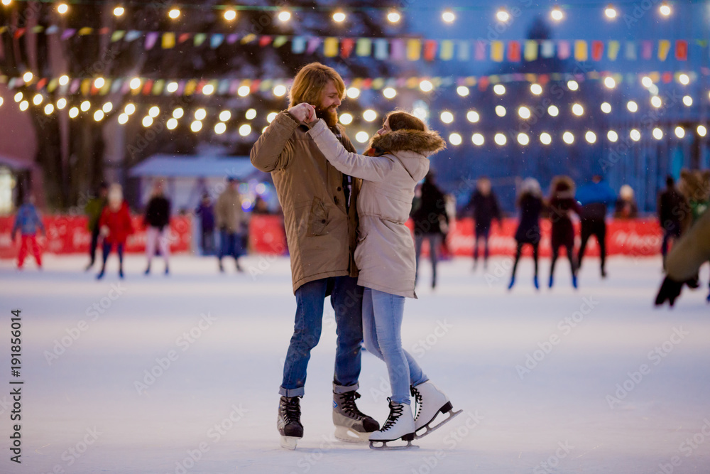 Theme ice skating rink and loving couple. meeting young, stylish people ride by hand in crowd on city skating rink lit by light bulbs and lights. Ice skating in winter for Christmas on ice arena - obrazy, fototapety, plakaty 