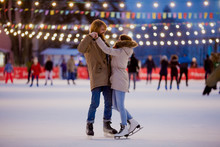 Theme Ice Skating Rink And Loving Couple. Meeting Young, Stylish People Ride By Hand In Crowd On City Skating Rink Lit By Light Bulbs And Lights. Ice Skating In Winter For Christmas On Ice Arena