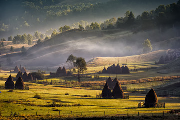 Beautiful rural mountain landscape in the morning light with fog, old houses and haystacks, Fundatura Ponorului, Hunedoara County, Romania