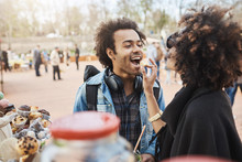 Side-view of cute african-american couple in love having fun in park during food festival, standing near counter and picking something to eat. Girlfriend feeds guy with sweet candy