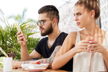 Wall Mural - Displeased jealous female sits back to her boyfriend who messages on smart phone and doesn`t pay attention, ignores girlfriend, spend free time together at outdoor terrace cafe. People and relations