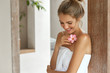 © wayhome.studio  - Beautiful young woman with soft skin, has light hair tied in knot, wears white towel after taking douche, smells flower, looks down with happy expression, pleased after good relax at bathroom