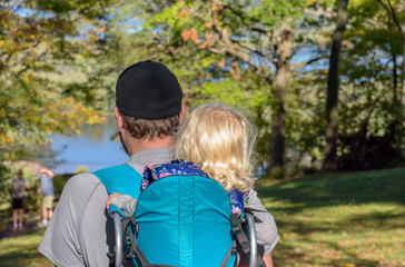 little girl sitting in backpack on dads back hiking in woods toward lake