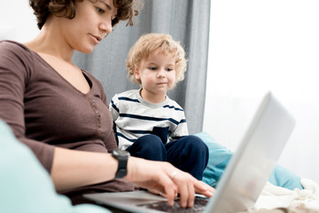 portrait of young mother using laptop at home, focus on curious little boy looking at screen and lea