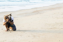 Man Sitting Alone With Hipster Hiker Backpack And Holding Camera Take Photo On The Beach With Tropical Sea Background. Concept Lifestyle Of Travel Photography. With Copy Space For Text.