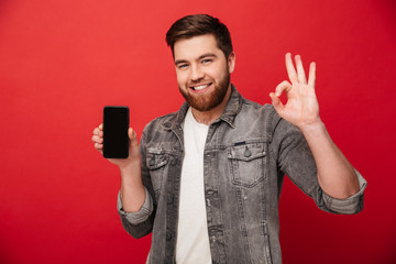 Wall Mural - Image of unshaved caucasian guy showing cell phone on camera and gesturing OK sign with fingers, isolated over red background