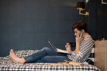 Side View  Portrait Of Teenage Boy Doing Homework While Lying On Bed, Using Laptop And Speaking By Phone At Same Time