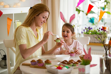 Wall Mural - Mother and daughter celebrating Easter, cooking cupcakes, covering with glaze. Happy family holiday. Cute little girl in bunny ears.