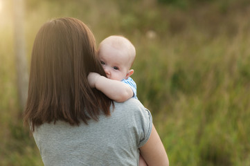 Wall Mural - Young tender brunette woman standing at green grass background hugging little cute child baby boy on nature. Mother, little kid son. Parenthood, family day 15 of may, love, parents, children concept.