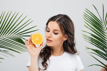 Wall Mural - Beauty portrait of a smiling woman with brunette hair posing with green leaves and holding sliced orange isolated over white background