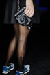 © Nikolay - A girl in a skirt is holding a professional camera in her hand. Leather belt. Blurred legs with tattoos in tights. Dark background