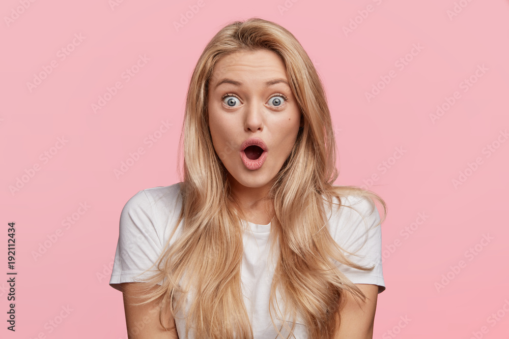 Blue eyed blonde young woman in casual white t shirt, looks with opened mouth, being shocked to recieve award after participating in competition, isolated over pink background. Shock concept - obrazy, fototapety, plakaty 