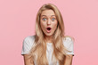 Blue eyed blonde young woman in casual white t shirt, looks with opened mouth, being shocked to recieve award after participating in competition, isolated over pink background. Shock concept