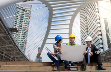 Picture of group professional Asian engineering team wearing safety helmet talking about blueprint looking paper plans at construction site with building city background