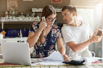 Wall Mural - Female in eyewear reads important documents and controls finances, speaks via smart phone with client, works on laptop computer and her fashionable husband, manage finances, count family budget