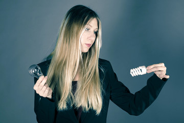 Wall Mural - Serious pretty business woman holding a light bulb on a grey background. Model looks at two kinds of lamps. Concept of choosing energy-saving light bulbs