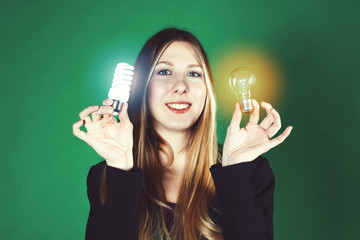 Wall Mural - Serious pretty business woman holding a light bulb on a green background. Close-up face model with two kinds of light bulbs. Concept of saving money for electricity