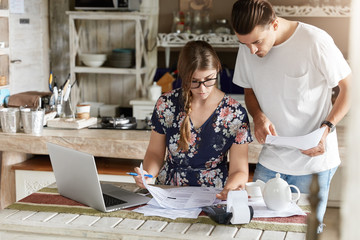 Pleasant looking housewife in glasses, makes necessary calculations, writes down information with pen, sits at kitchen table with documents, calculator and laptop computer, young man helps wife