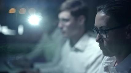 Concentrated female cyber security professional working on computer at night with male colleagues; projector lightning at camera in background