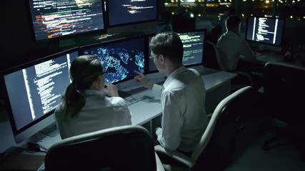 Tilt up of man and woman sitting in dark office at night and discussing program code and holographic world map on multiple monitors