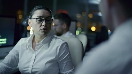 Tracking of female IT programmer working on computer and then turning to male colleague and speaking with him while working in dark office at night