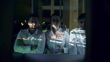 Zoom in of team of cyber security professionals standing in dark office at night and discussing programming code; computer data projected on his face
