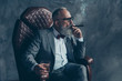 © deagreez - Side profile view of  thoughtful, serious, minded, ponder rich man in glasses, smoke cigarette, fume, hold glass with brandy, in tux with red bow tie, sit in leather armchair over gray background