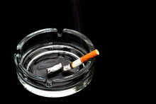 Smoldering Cigarette In A Glassy Ashtray Isolated On Black Background