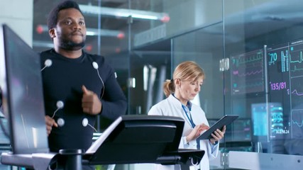 Wall Mural - male athlete runs on a treadmill with electrodes attached to his body while sport scientist holds ta