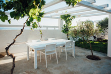 Terrace With White Table And Chairs Under A White Roof With Vine And Olive Trees On The Island Of Santorini