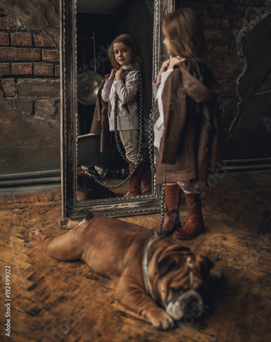 Fotomural Child girl in image of Sherlock Holmes stands next to English bulldog and looks at mirror reflection on background of old interior