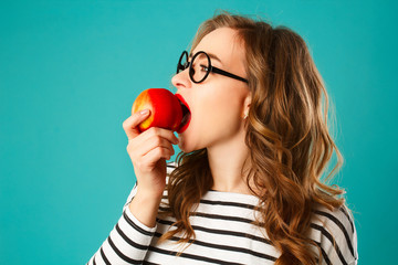 Wall Mural - Portrait of young beautiful blond woman in round black glasses eating red apple over blue background