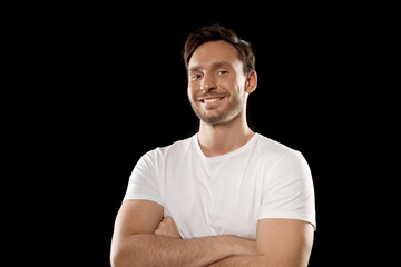 Handsome young man in a white t shirt isolated on a black background. Happy guy is smiling with his hands crossed