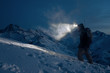 Professional expeditor commit climb on snowy mountains at night and lights the way with a headlamp. Wearing ski wear, backpack and a snowboard behind his back. Backcountry and ski touring