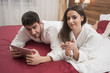 © Senad - Beautiful happy young couple enjoying in bed after spa. Couple in white robes relaxing. Man using tablet while having conversation with woman