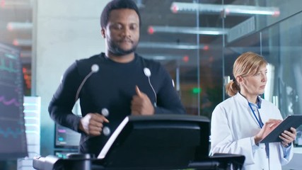 Wall Mural - male athlete runs on a treadmill with electrodes attached to his body while sport scientist holds ta