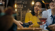 © Gorodenkoff - In the Bar Waiter Holds Credit Card Payment Machine and Beautiful Woman Pays for Her Order with Contactless Mobile Phone Payments System. She's Surrounded by Dear Friends and Has Time of Her Life.