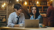 © Gorodenkoff - Handsome Asian Man and Beautiful Caucasian Young Woman Sitting in the Cafe Work on a Laptop Computer. In the Background Other Customers in the Stylish Environment.
