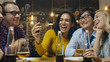 © Gorodenkoff - In the Bar/ Restaurant Hispanic Woman Makes Video Call with Her Friends. Group Beautiful Young People in Stylish Establishment.
