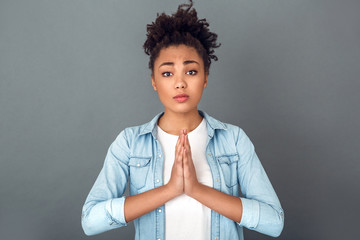 Wall Mural - Young african woman isolated on grey wall studio casual daily lifestyle praying gesture