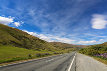 A Straight Country Road Between Green Hills In New Zealand. Bright Blue Sky With White Clouds, Sunny Day