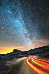 Wall Mural - Night landscape. Night sky with a north hemisphere Milky Way and stars. The night road illuminated by the car winds with a serpentine and leaves in a distance to a foot of an acute rock. Light trails