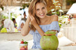 © WHstudio Leushin N - Beautiful happy woman with positive expression, being glad to meet with best friend in cafeteria, drinks coconut cocktail and eats dessert, sits against cozy interior. People, happiness and resort